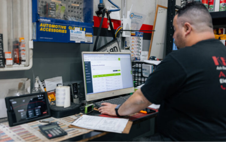 A man in a black t-shirt sits at a desk in an automotive shop, working on a computer that displays a "Roadworthy eCertificate" portal. The desk is cluttered with a tablet, a calculator, and paperwork, with shelves of automotive parts and accessories visible in the background.