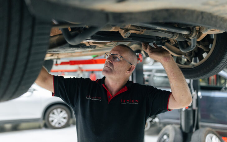 A mechanic in a black and red polo shirt inspects the undercarriage of a car lifted on a hydraulic hoist.